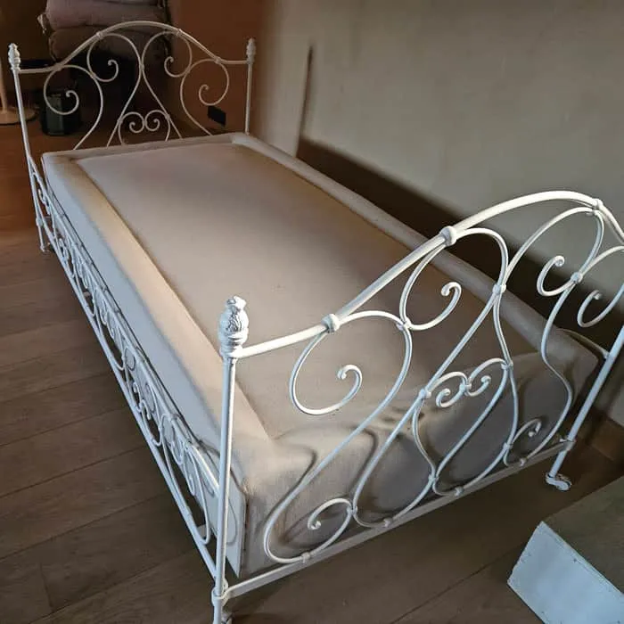 Lit ancien en fer forgé blanc posé sur parquet, sommier et matelas beige empilés